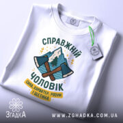 Футболка справжній чоловік, біла, зображена зверху, на фоні світло-сірого кольору, принт з двома сокирами та горами, текст українською мовою, з етикеткою на комірці.