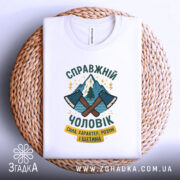 Футболка справжній чоловік, біла, зображена зверху, на фоні плетеного кошика, принт з двома сокирами та горами, текст українською мовою.