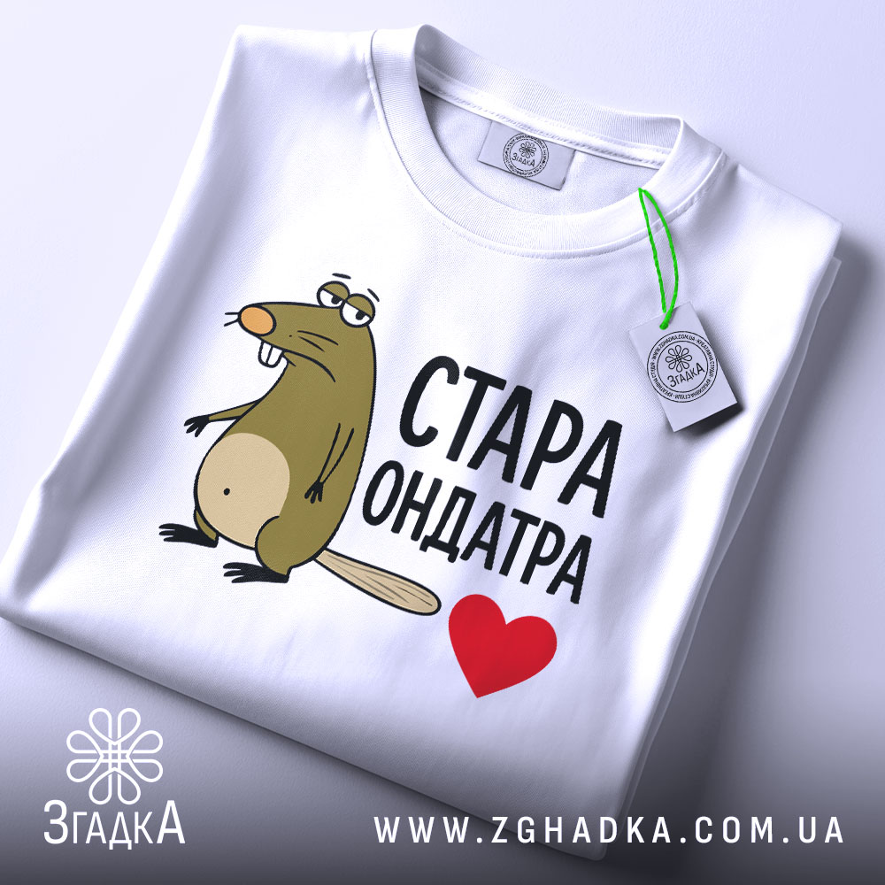 Футболка стара ондатра, білого кольору, зображена зверху, складена на поверхні, фон світло-сірий, принт з мультяшною ондатрою та написом 'СТАРА ОНДАТРА' чорного кольору з червоним сердечком, на футболці є етикетка з зеленим шнурком.