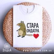Футболка стара ондатра, білого кольору, зображена зверху, складена на плетеній основі, фон світло-сірий, принт з мультяшною ондатрою та написом 'СТАРА ОНДАТРА' чорного кольору з червоним сердечком.