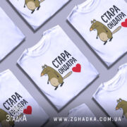 Футболка стара ондатра, білого кольору, зображена з правого ракурсу на манекені, фон світло-сірий, принт з мультяшною ондатрою та написом 'СТАРА ОНДАТРА' чорного кольору з червоним сердечком.