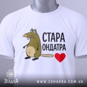 Футболка стара ондатра, білого кольору, зображена зверху, складена на поверхні, фон світло-сірий, принт з мультяшною ондатрою та написом 'СТАРА ОНДАТРА' чорного кольору з червоним сердечком, на футболці є етикетка з зеленим шнурком.