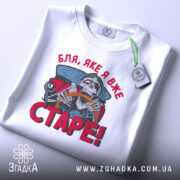Футболка старе скелет, біла, складена на поверхні, фон світло-сірий, принт з черепом за кермом автомобіля, текст червоним кольором 'Бля, яке я вже СТАРЕ!', на етикетці зелена стрічка.