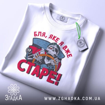Футболка старе скелет, біла, складена на поверхні, фон світло-сірий, принт з черепом за кермом автомобіля, текст червоним кольором 'Бля, яке я вже СТАРЕ!', на етикетці зелена стрічка.