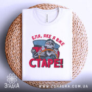 Футболка старе скелет, біла, складена на плетеній кошику, фон світло-сірий, принт з черепом за кермом автомобіля, текст червоним кольором 'Бля, яке я вже СТАРЕ!'.