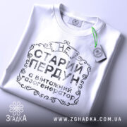 Футболка старий пердун, біла, складена на поверхні, фон світлий, принт чорного кольору з декоративними елементами, на ярлику зелена стрічка.