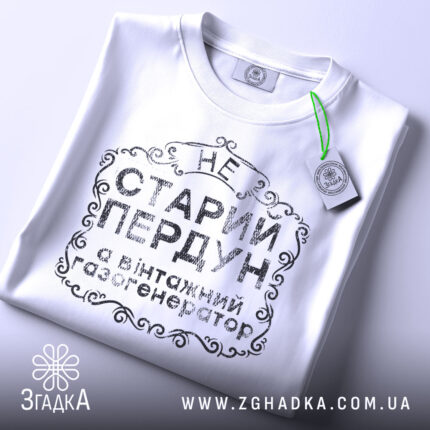 Футболка старий пердун, біла, складена на поверхні, фон світлий, принт чорного кольору з декоративними елементами, на ярлику зелена стрічка.