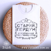 Футболка старий пердун, біла, складена на плетеній тарілці, фон світлий, принт чорного кольору з декоративними елементами.