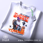 Футболка старий пердун. Біла футболка, складена на столі, з видимим етикеткою. Фон світло-сірий, з легким відблиском. Принт з дідусем у фіолетовій шапці, текст червоний і жовтий, зображення яскраве і детальне.