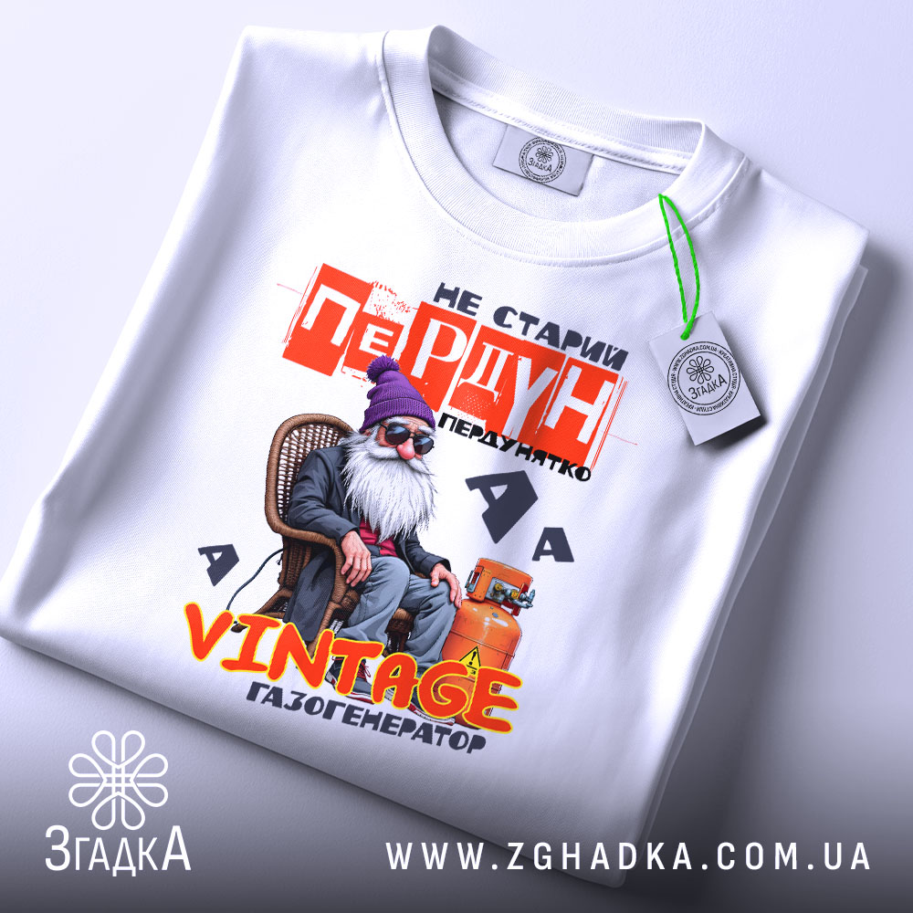 Футболка старий пердун. Біла футболка, складена на столі, з видимим етикеткою. Фон світло-сірий, з легким відблиском. Принт з дідусем у фіолетовій шапці, текст червоний і жовтий, зображення яскраве і детальне.