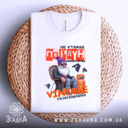 Футболка старий пердун. Біла футболка, складена на плетеній основі, фон білий. Принт з дідусем у фіолетовій шапці, текст червоний і жовтий, виглядає стильно і оригінально.