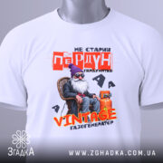 Футболка старий пердун. Біла футболка, складена на столі, з видимим етикеткою. Фон світло-сірий, з легким відблиском. Принт з дідусем у фіолетовій шапці, текст червоний і жовтий, зображення яскраве і детальне.