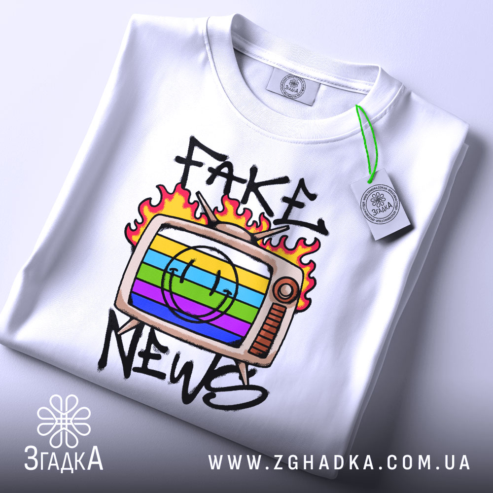Футболка стоп фейкам, складена біла футболка з принтом телевізора, на якому зображено кольорові смужки та напис 'FAKE NEWS', етикетка з зеленим шнурком, фон світлий.