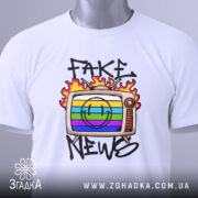 Футболка стоп фейкам, складена біла футболка з принтом телевізора, на якому зображено кольорові смужки та напис 'FAKE NEWS', етикетка з зеленим шнурком, фон світлий.