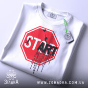 Футболка стоп старт арт, біла, складена на поверхні, фон світлий, принт червоного кольору у формі знака стоп з написом 'START' чорним кольором, з елементами графіті, на ярлику зелена стрічка.