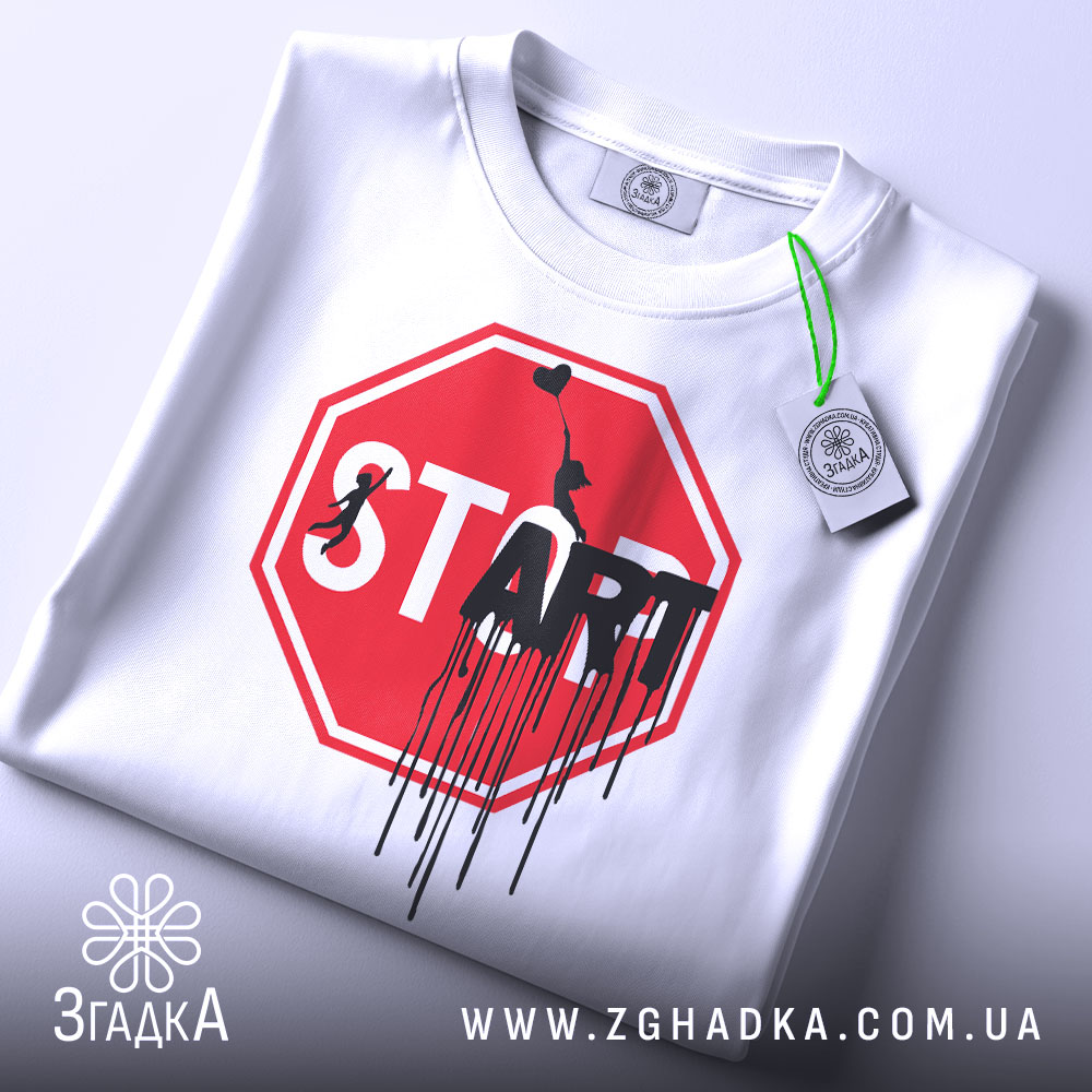 Футболка стоп старт арт, біла, складена на поверхні, фон світлий, принт червоного кольору у формі знака стоп з написом 'START' чорним кольором, з елементами графіті, на ярлику зелена стрічка.