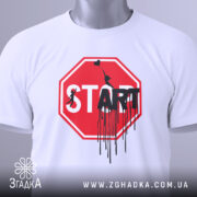 Футболка стоп старт арт, біла, складена на поверхні, фон світлий, принт червоного кольору у формі знака стоп з написом 'START' чорним кольором, з елементами графіті, на ярлику зелена стрічка.