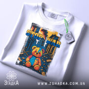 Футболка стоп війна, біла, складена на поверхні, фон світлий, принт з ведмедиком, прикріплена етикетка з зеленим шнурком.