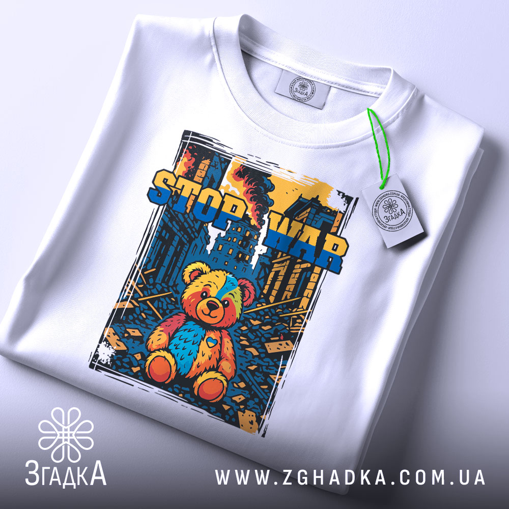 Футболка стоп війна, біла, складена на поверхні, фон світлий, принт з ведмедиком, прикріплена етикетка з зеленим шнурком.