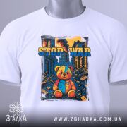 Футболка стоп війна, біла, складена на поверхні, фон світлий, принт з ведмедиком, прикріплена етикетка з зеленим шнурком.