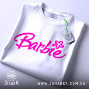 Футболка стиль барбі, біла, складена на поверхні, фон світлий, принт з написом 'Barbie' яскравого рожевого кольору, з етикеткою на комірці.