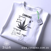 Футболка стиль канабіс, біла, складена на поверхні, фон світло-сірий, принт чорного кольору з малюнком листа канабісу та текстом, з зеленим ярликом.