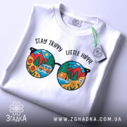 Футболка стиль хіпі, біла, складена на поверхні, фон світло-сірий, принт у вигляді кольорових окулярів з пейзажем, текст 'STAY TRIPPY LITTLE HIPPIE' над окулярами, на ярлику зелена стрічка.