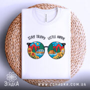 Футболка стиль хіпі, біла, складена на плетеній кошику, фон світло-сірий, принт у вигляді кольорових окулярів з пейзажем, текст 'STAY TRIPPY LITTLE HIPPIE' над окулярами.