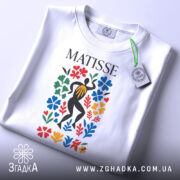 Футболка стиль Матісса, біла, складена на столі, фон світло-сірий, принт з кольоровими квітами та фігурою людини в чорному, з етикеткою.