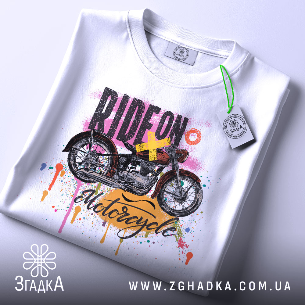 Футболка стиль мотоцикліста, біла, складена на поверхні, з видимим етикеткою, фон світлий, принт з мотоциклом та кольоровими краплями фарби.