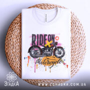 Футболка стиль мотоцикліста, біла, складена на плетеній кошику, фон світлий, принт з мотоциклом та кольоровими краплями фарби.