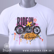 Футболка стиль мотоцикліста, біла, складена на поверхні, з видимим етикеткою, фон світлий, принт з мотоциклом та кольоровими краплями фарби.
