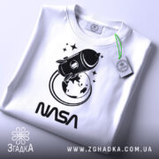 Футболка стиль NASA, біла, складена на столі, фон світло-сірий, принт з ракетою та Землею в чорному кольорі, текст 'NASA' під принтом, на футболці є етикетка.