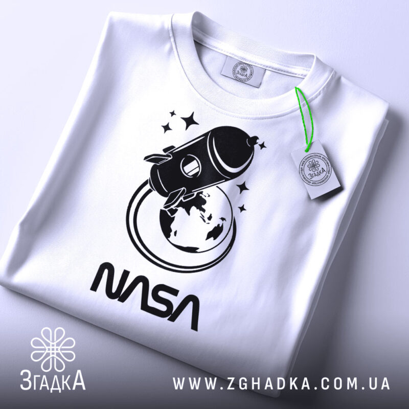 Футболка стиль NASA, біла, складена на столі, фон світло-сірий, принт з ракетою та Землею в чорному кольорі, текст 'NASA' під принтом, на футболці є етикетка.