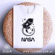 Футболка стиль NASA, біла, складена на плетеній кошику, фон світло-сірий, принт з ракетою та Землею в чорному кольорі, текст 'NASA' під принтом.