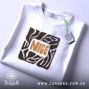 Футболка стиль NICE, біла, складена на столі, фон світло-сірий, принт з геометричним малюнком та написом 'NICE' оранжевого кольору, на футболці є етикетка з зеленим шнурком.