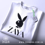 Футболка стиль зая білого кольору, складена на поверхні, зображена з верхнього ракурсу, фон світло-сірий, принт чорного кольору з логотипом ZAYA та зображенням зайця в смокінгу, на футболці є етикетка з зеленим шнурком.