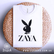 Футболка стиль зая білого кольору, складена на плетеній кошику, зображена з верхнього ракурсу, фон світло-сірий, принт чорного кольору з логотипом ZAYA та зображенням зайця в смокінгу.