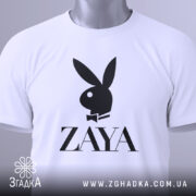 Футболка стиль зая білого кольору, складена на поверхні, зображена з верхнього ракурсу, фон світло-сірий, принт чорного кольору з логотипом ZAYA та зображенням зайця в смокінгу, на футболці є етикетка з зеленим шнурком.