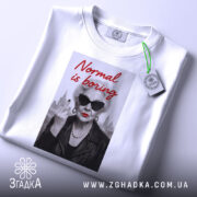 Футболка стильна бабуся, біла, з верхнього ракурсу, на фоні світло-сірого кольору, принт з зображенням бабусі в чорних окулярах, з червоними губами, в шкірянці, з написом 'Normal is boring' червоним шрифтом, з етикеткою зеленого кольору.