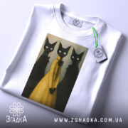 Футболка стильні коти арт, складена біла футболка на світлому фоні, принт з трьома чорними котами, один з яких у жовтій сукні, на футболці є етикетка з зеленим шнурком.