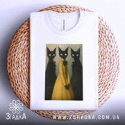 Футболка стильні коти арт, складена біла футболка на плетеній кошику, фон світлий, принт з трьома чорними котами, один з яких у жовтій сукні, коти мають великі білі очі.
