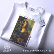 Футболка сучасна моналіза, білого кольору, зображена в плоскому вигляді, фон світлий, принт з Моною Лізою з короною, прикріплена етикетка з зеленим шнурком, текст на принті читається.