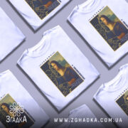 Футболка сучасна моналіза, білого кольору, зображена на манекені, фон нейтральний, принт зображає стилізовану версію Мони Лізи з короною, деталі принта включають текст і графічні елементи.