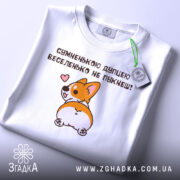 Футболка сумненькою дупцею не пукнеш. Біла футболка, складена на столі з легким блиском. Принт корги з сердечком на фоні світлого кольору, з етикеткою, що звисає з коміра.