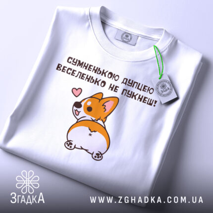 Футболка сумненькою дупцею не пукнеш. Біла футболка, складена на столі з легким блиском. Принт корги з сердечком на фоні світлого кольору, з етикеткою, що звисає з коміра.