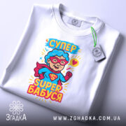 Футболка супер бабуся, білого кольору, складена на поверхні, фон світло-сірий, принт з персонажем у червоному плащі та окулярах, текст 'СУПЕР БАБУСЯ' яскравими кольорами, з прикріпленою етикеткою.