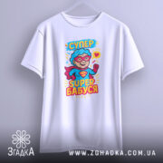 Футболка супер бабуся, білого кольору, підвішена на вішалці, фон світло-сірий, принт з персонажем у червоному плащі та окулярах, текст 'СУПЕР БАБУСЯ' яскравими кольорами.