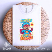 Футболка супер бабуся, білого кольору, складена на кошику з плетеного матеріалу, фон білий, принт з персонажем у червоному плащі та окулярах, текст 'СУПЕР БАБУСЯ' яскравими кольорами.