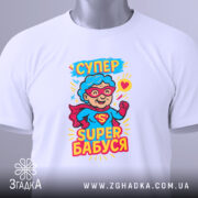 Футболка супер бабуся, білого кольору, складена на поверхні, фон світло-сірий, принт з персонажем у червоному плащі та окулярах, текст 'СУПЕР БАБУСЯ' яскравими кольорами, з прикріпленою етикеткою.